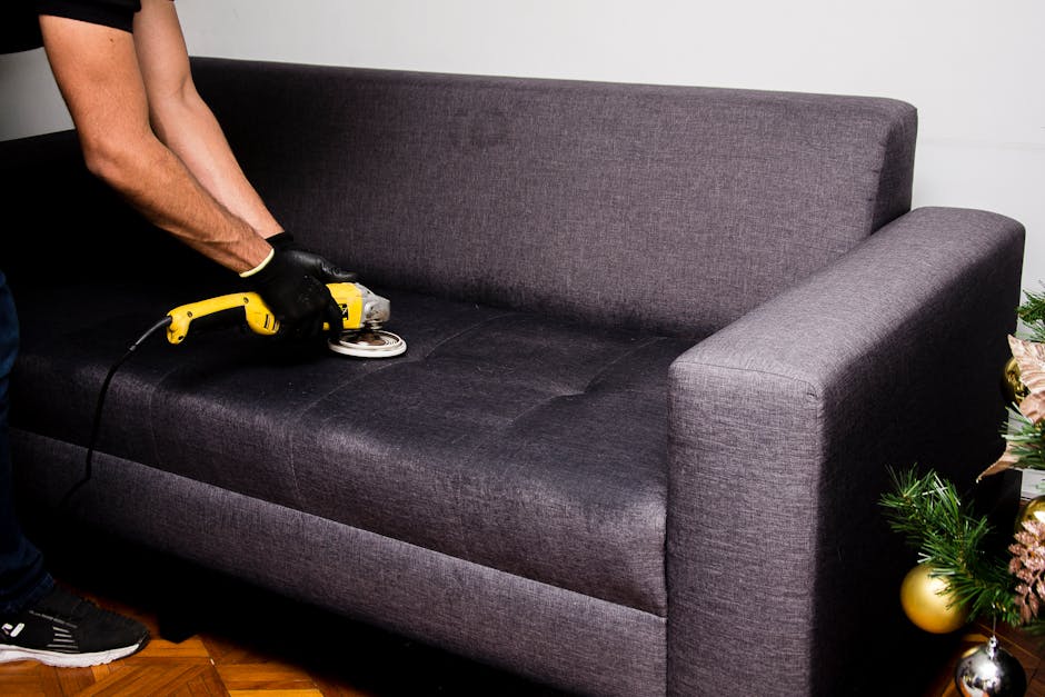 A person wearing black gloves is using a handheld electric upholstery cleaner to deep clean a dark gray fabric sofa in a well-lit room. The sofa has a modern design with a straight back and wide armre
