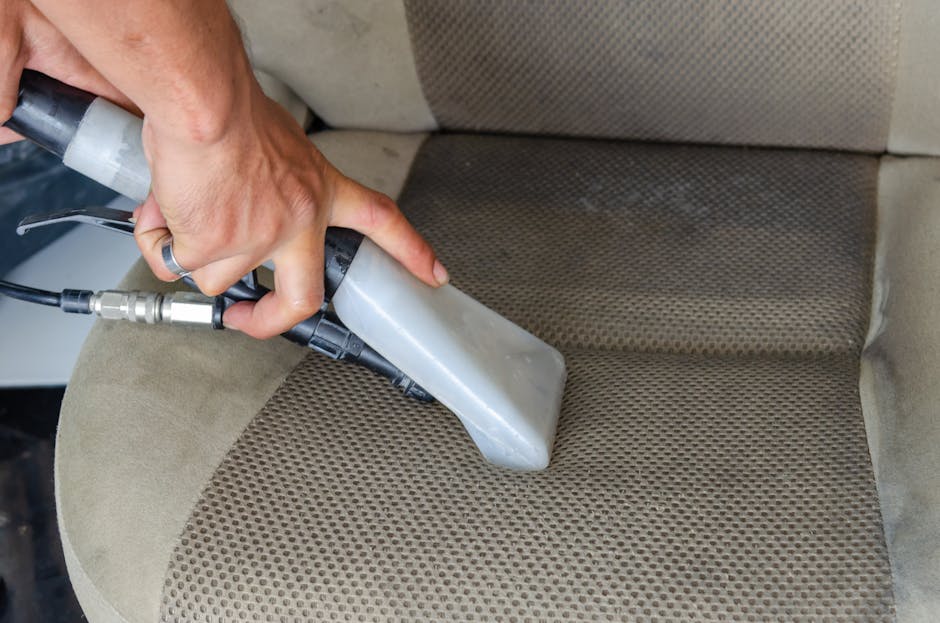 A person wearing black gloves is using a handheld electric upholstery cleaner to deep clean a dark gray fabric sofa in a well-lit room. The sofa has a modern design with a straight back and wide armre