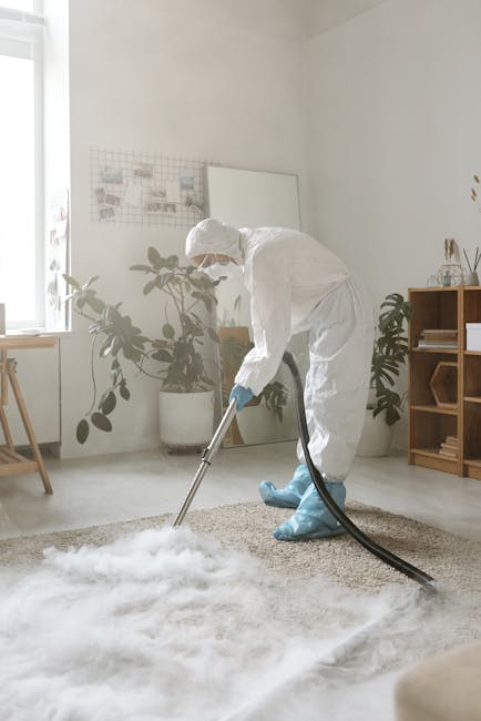 A person using a yellow vacuum cleaner with a black hose to deep clean a traditional floral-patterned rug in a residential living room. The rug features muted tones of beige, green, and pink, and is s